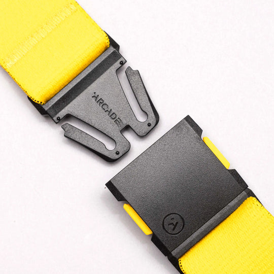 A close-up of a yellow National Geographic belt by Arcade Belts, featuring a black, partially unclasped low-profile buckle engraved with ARCADE, set against a plain white background.