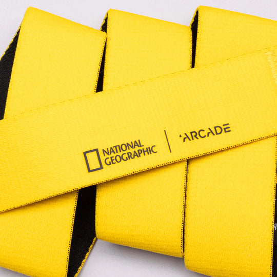 The National Geographic belt by Arcade Belts features black text and the National Geographic logo on bright yellow stretch webbing, artfully arranged in overlapping loops on a light background.