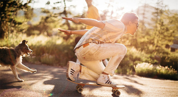 A person in sunglasses and light clothing, secured by a low profile belt, crouches on a skateboard with arms outstretched, riding down a sunlit path as a dog runs beside them. Trees and another person appear in the bright background.