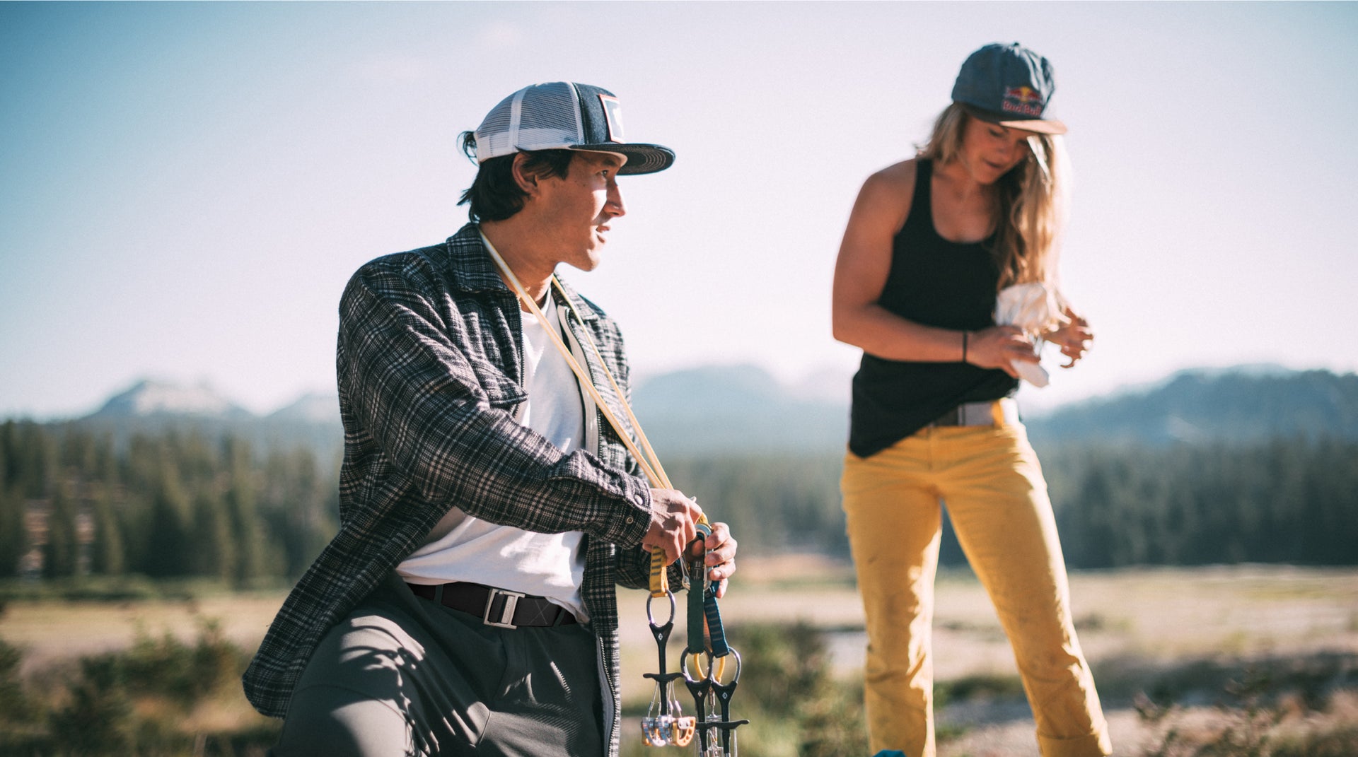 A man and woman, both wearing hats, stand outdoors in a scenic mountain landscape. The man holds climbing equipment and wears a flannel shirt, while the woman, in a tank top and yellow pants with a travel belt, looks down at her hands.