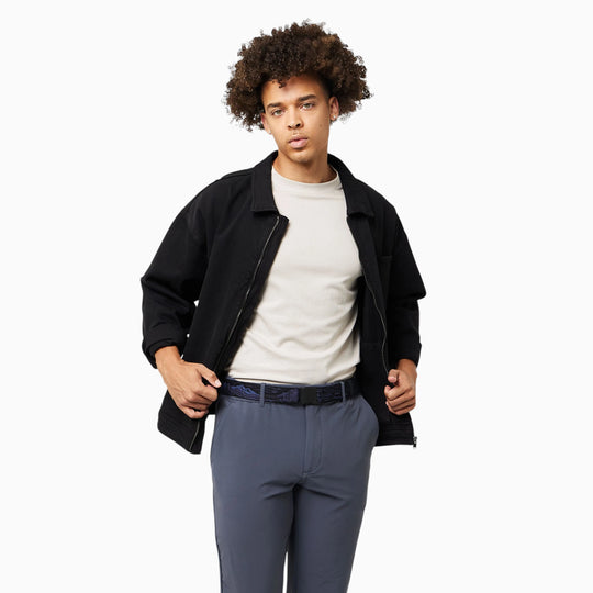 A young person with curly hair stands against a plain white background with a relaxed expression, wearing the Arcade Belts Out of Range Kit—a black jacket featuring performance stretch webbing—layered over a light shirt and paired with gray pants.