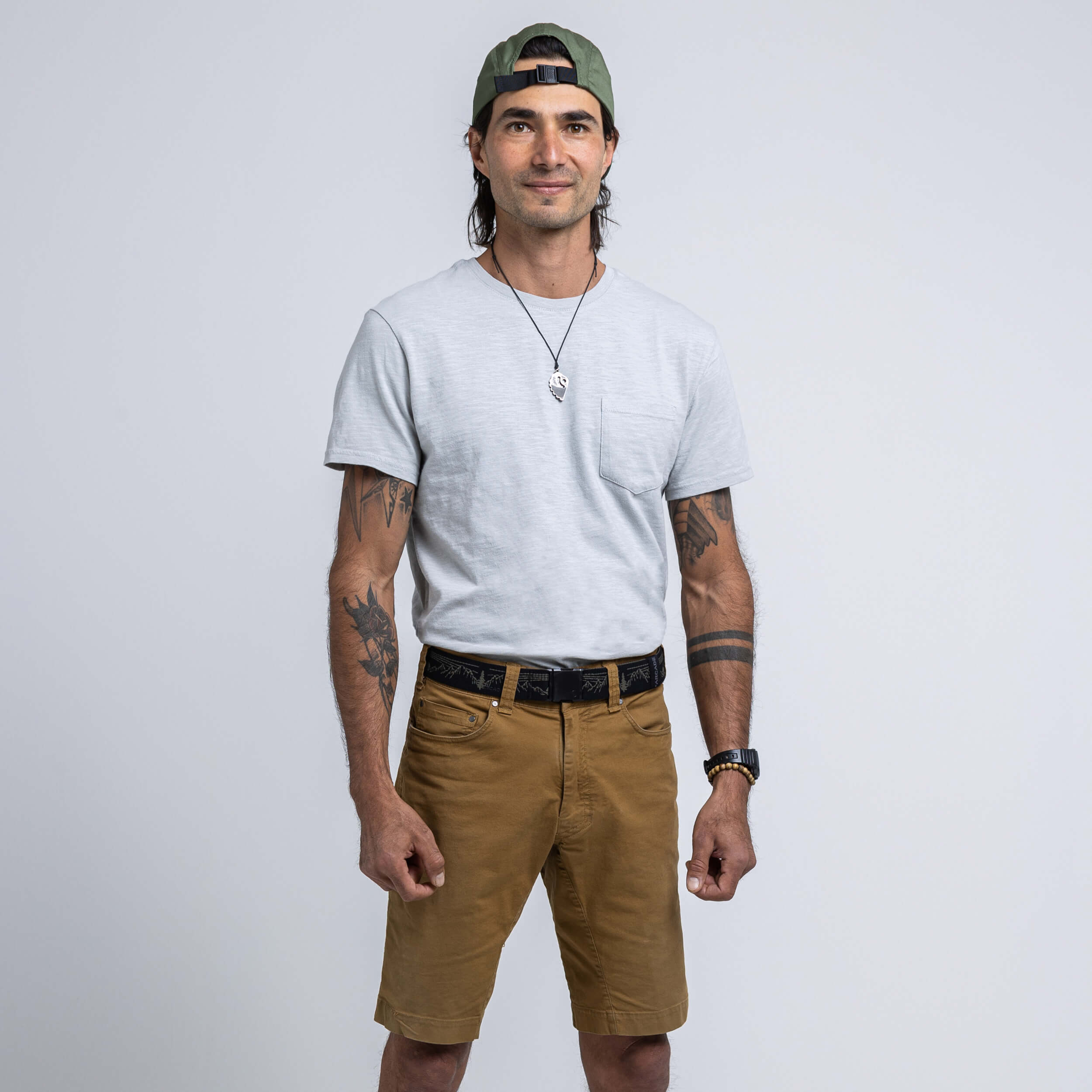 A man models the Arcade Belts Out of Range Kit, wearing a light grey T-shirt, brown performance stretch shorts, and a green backwards cap. He sports tattoos on both arms, a necklace, and a slight smile with his hands at his sides.