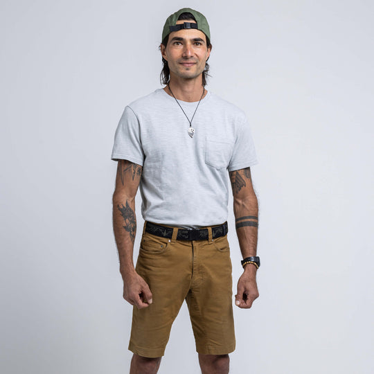 A man models the Arcade Belts Out of Range Kit, wearing a light grey T-shirt, brown performance stretch shorts, and a green backwards cap. He sports tattoos on both arms, a necklace, and a slight smile with his hands at his sides.