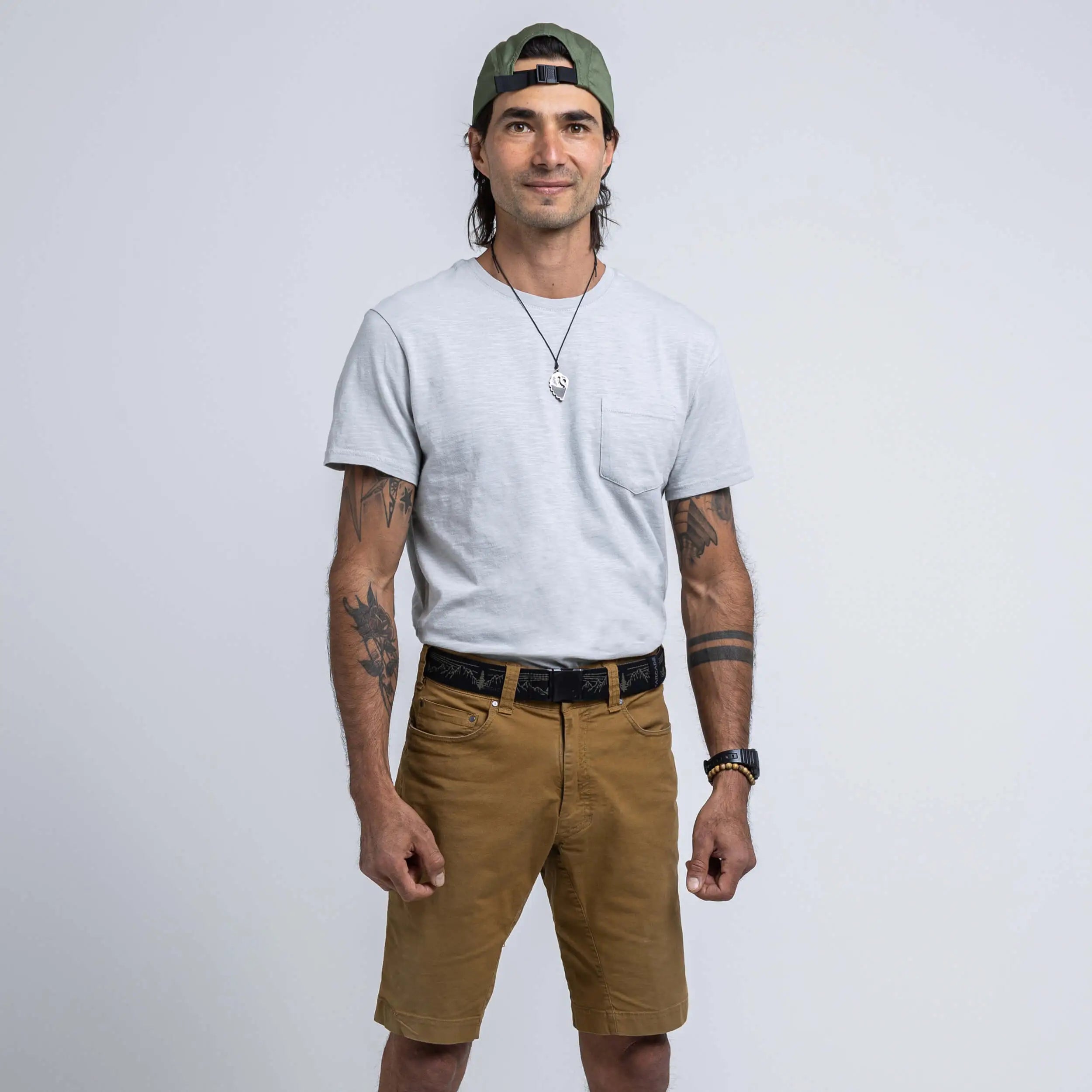 A man models the Arcade Belts Out of Range Kit, wearing a light grey T-shirt, brown performance stretch shorts, and a green backwards cap. He sports tattoos on both arms, a necklace, and a slight smile with his hands at his sides.
