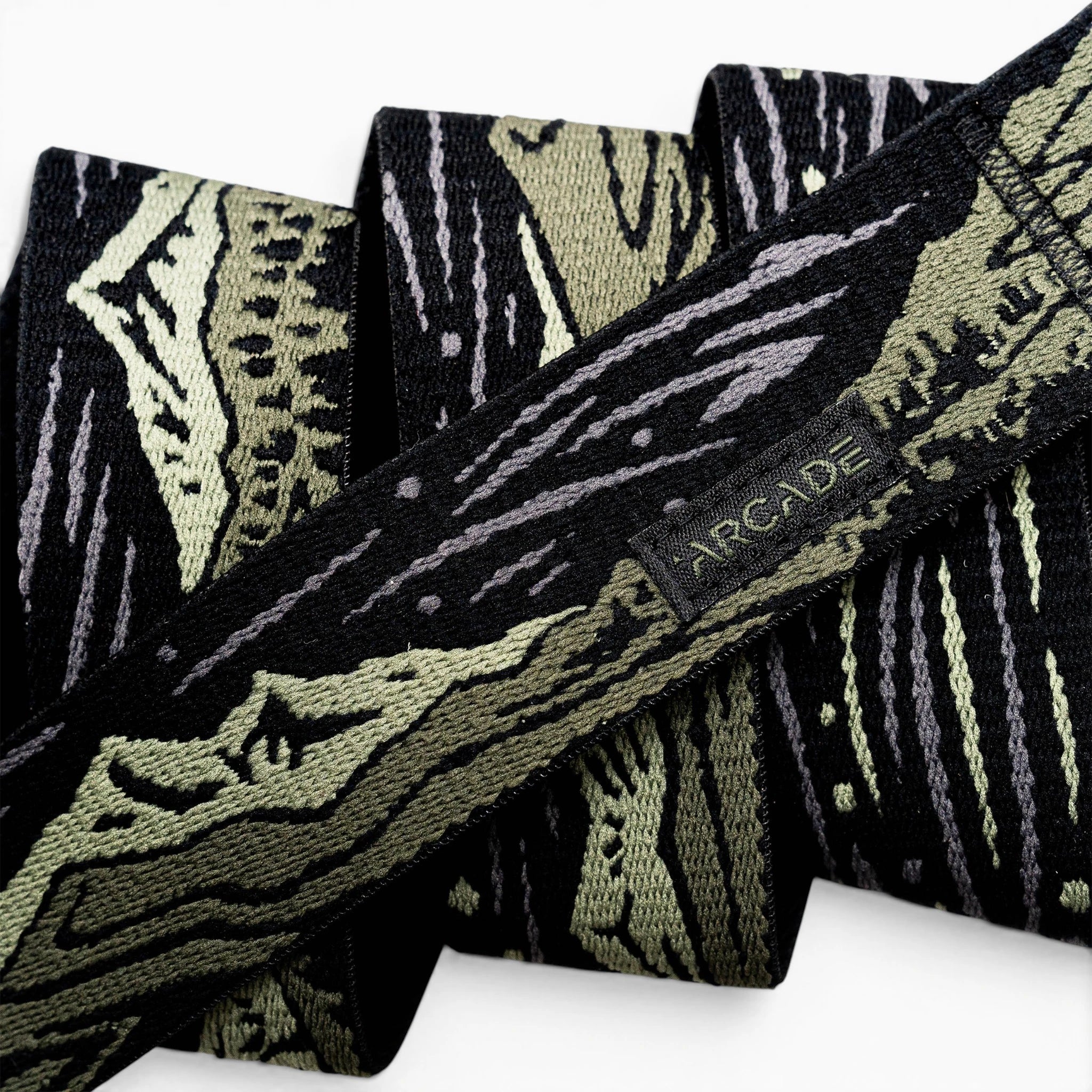 A close-up of the Arcade Belts Out of Range fabric belt, showcasing a black and olive green camo pattern, a small black tag with the brand name stitched on it, and a sturdy plastic buckle.