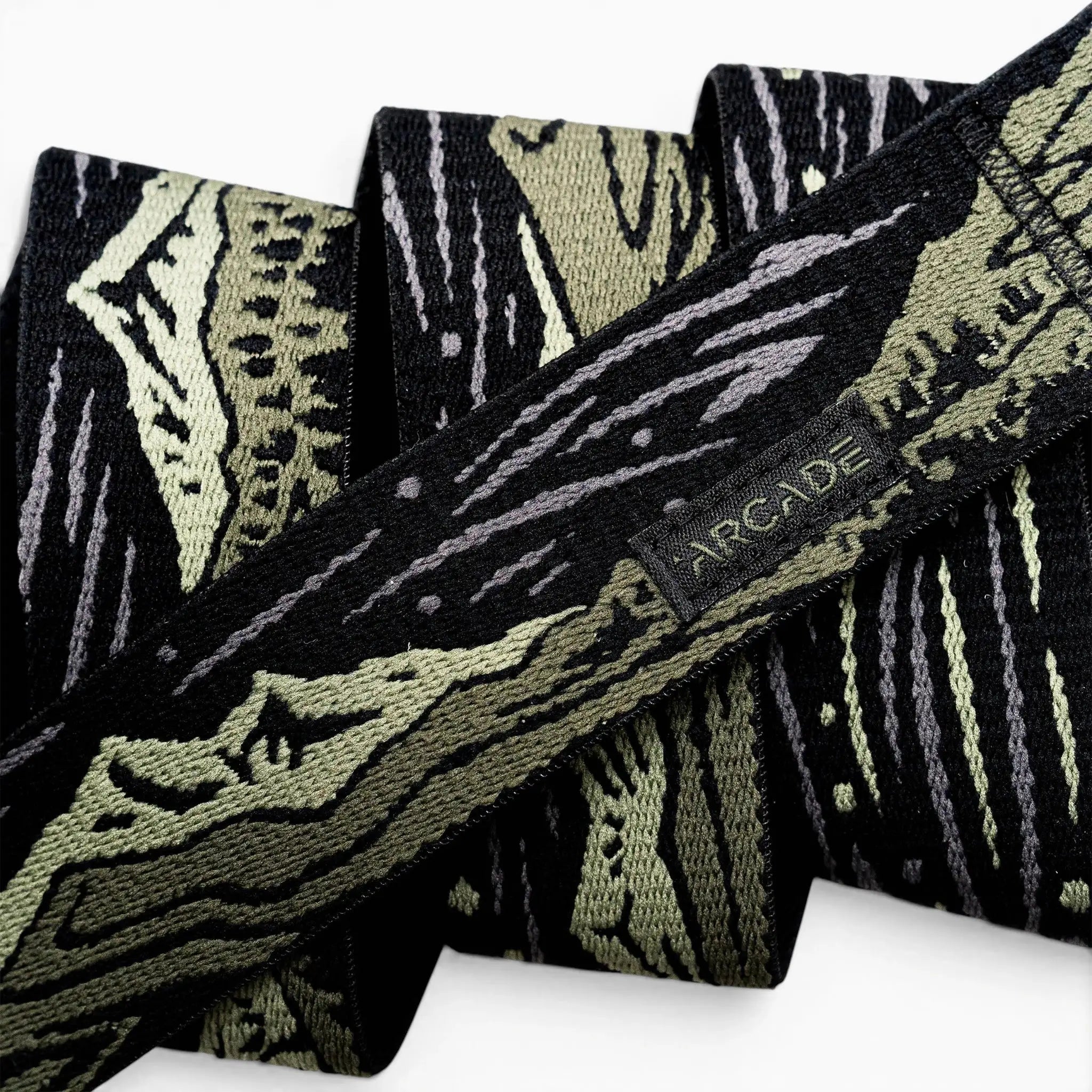 A close-up of the Arcade Belts Out of Range fabric belt, showcasing a black and olive green camo pattern, a small black tag with the brand name stitched on it, and a sturdy plastic buckle.