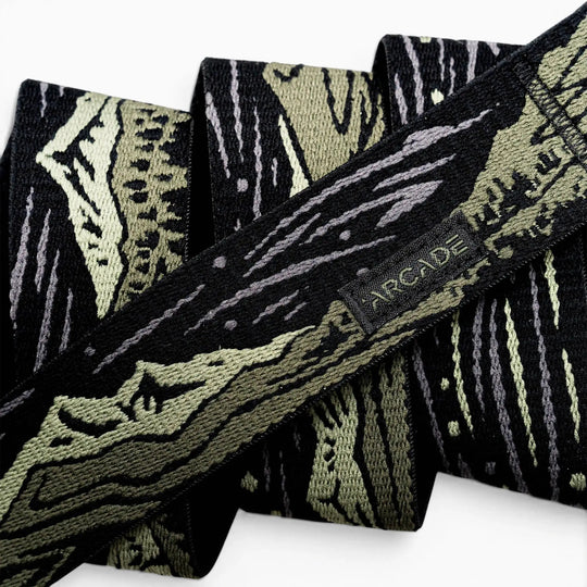 A close-up of the Arcade Belts Out of Range fabric belt, showcasing a black and olive green camo pattern, a small black tag with the brand name stitched on it, and a sturdy plastic buckle.