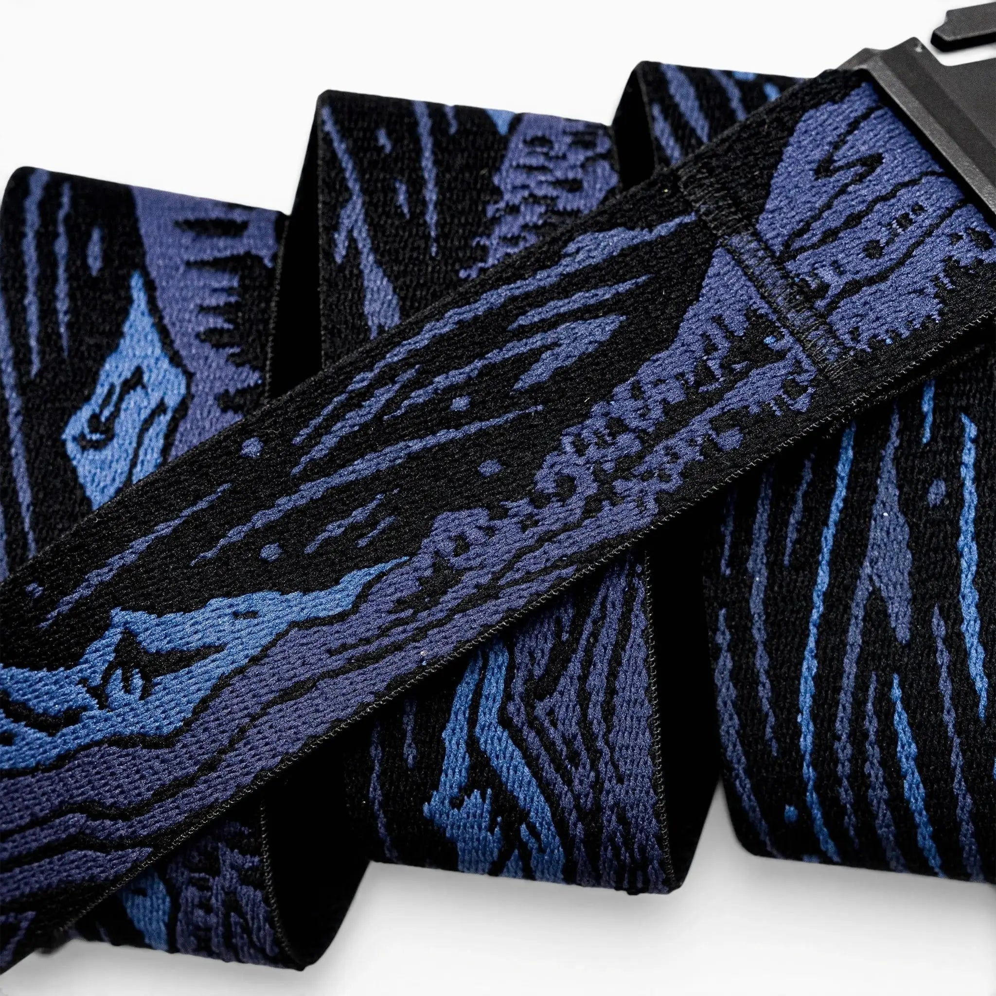 A close-up of the Arcade Belts Out of Range belt, showcasing its low profile design, black and blue abstract woodgrain pattern, and black buckle. The travel belt is neatly folded over itself multiple times.