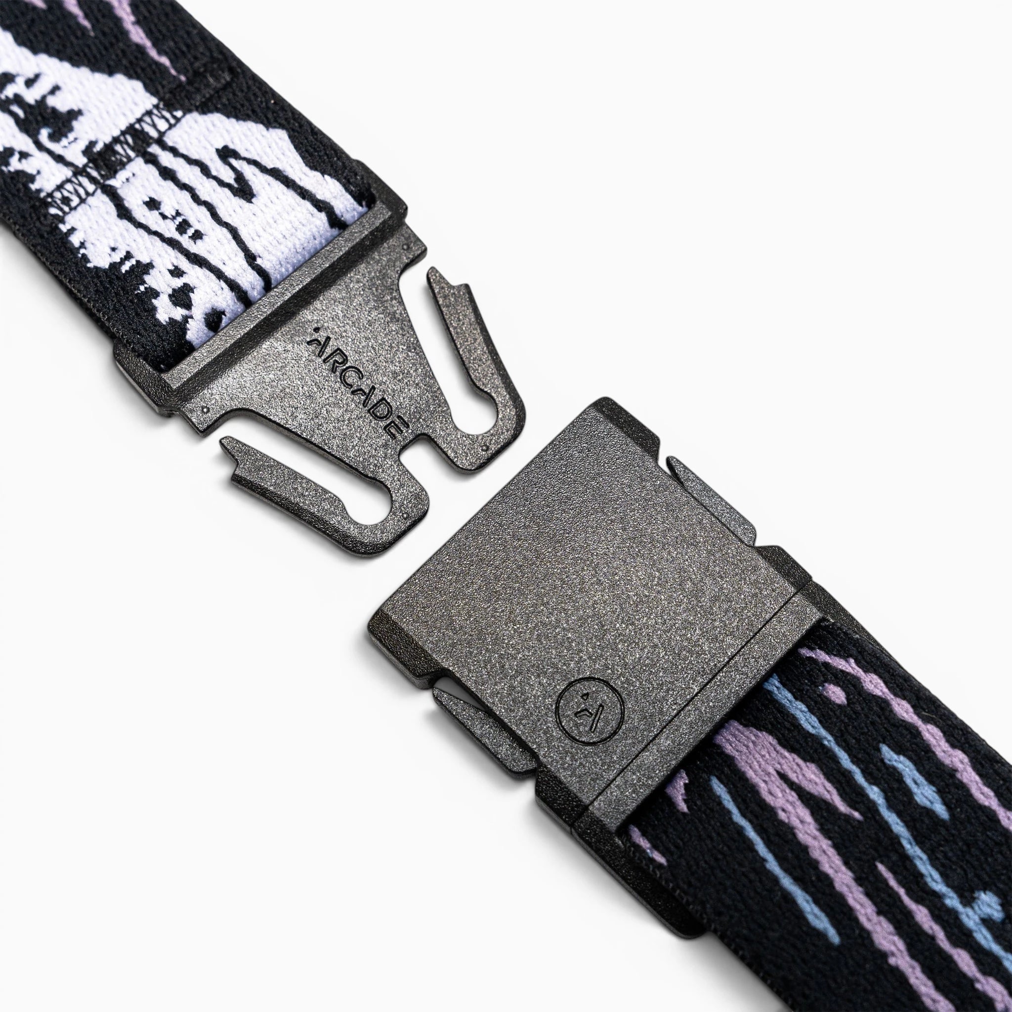 A close-up of the Arcade Belts Slim Kit shows its black plastic buckle on a stretch web strap with white, black, purple, and blue patterns, unclasped on a white background—perfect as a low-profile belt.