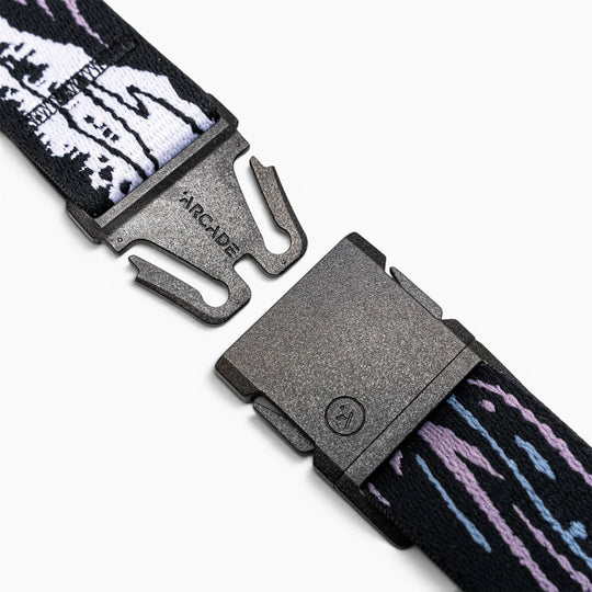 A close-up of the Arcade Belts Slim Kit shows its black plastic buckle on a stretch web strap with white, black, purple, and blue patterns, unclasped on a white background—perfect as a low-profile belt.
