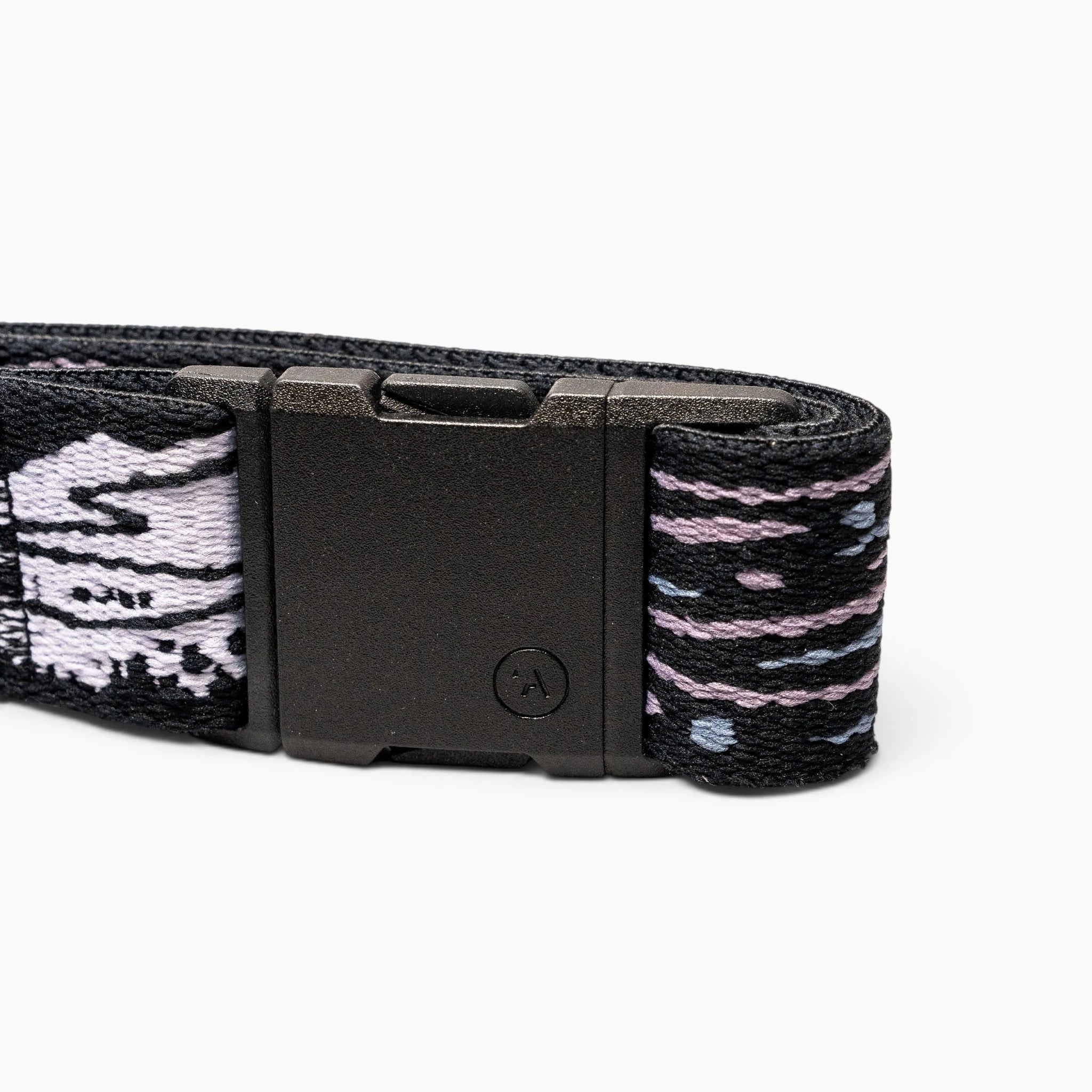 A close-up of the Arcade Belts Out of Range black elastic stretch belt with a black rectangular buckle and patterned fabric in black, white, and purple, coiled on a white background.