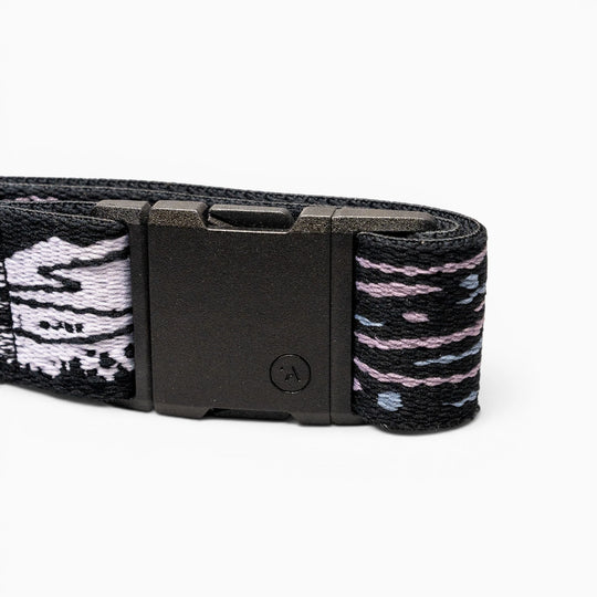 A close-up of the Arcade Belts Out of Range black elastic stretch belt with a black rectangular buckle and patterned fabric in black, white, and purple, coiled on a white background.