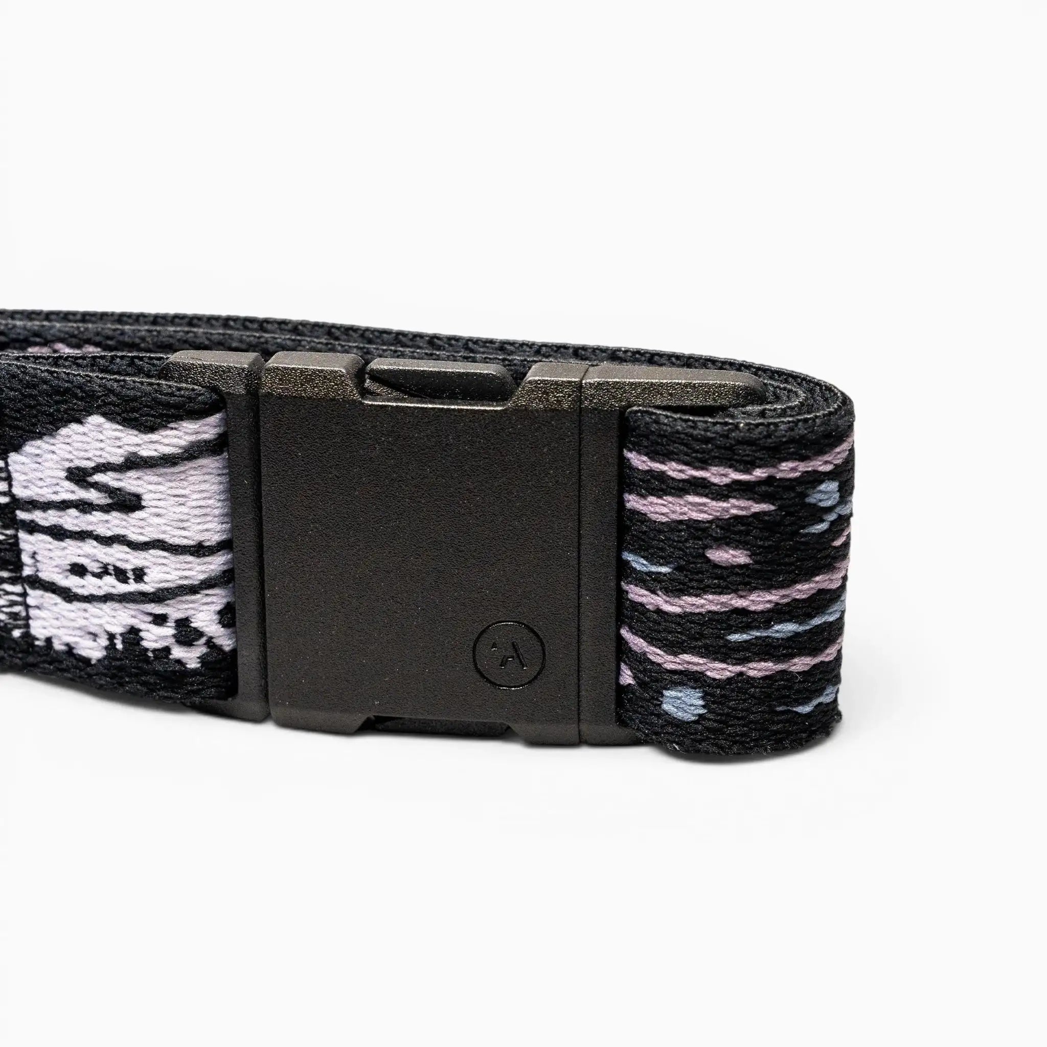A close-up of the Arcade Belts Out of Range black elastic stretch belt with a black rectangular buckle and patterned fabric in black, white, and purple, coiled on a white background.