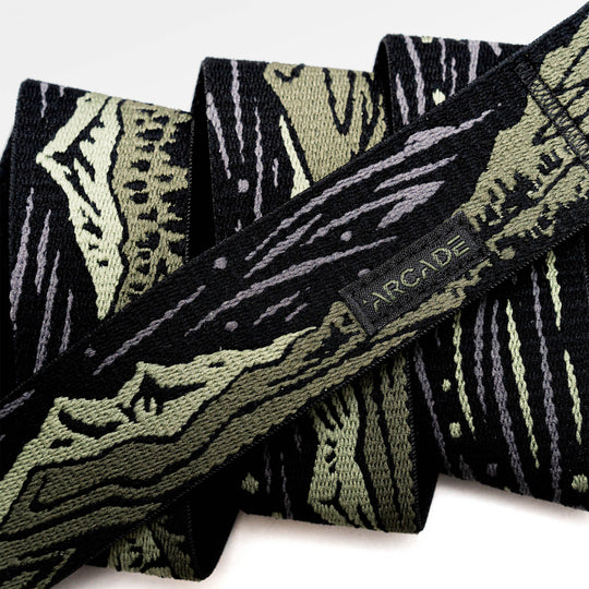 A close-up of the Arcade Belts Out of Range fabric belt, showcasing a black and olive green camo pattern, a small black tag with the brand name stitched on it, and a sturdy plastic buckle.
