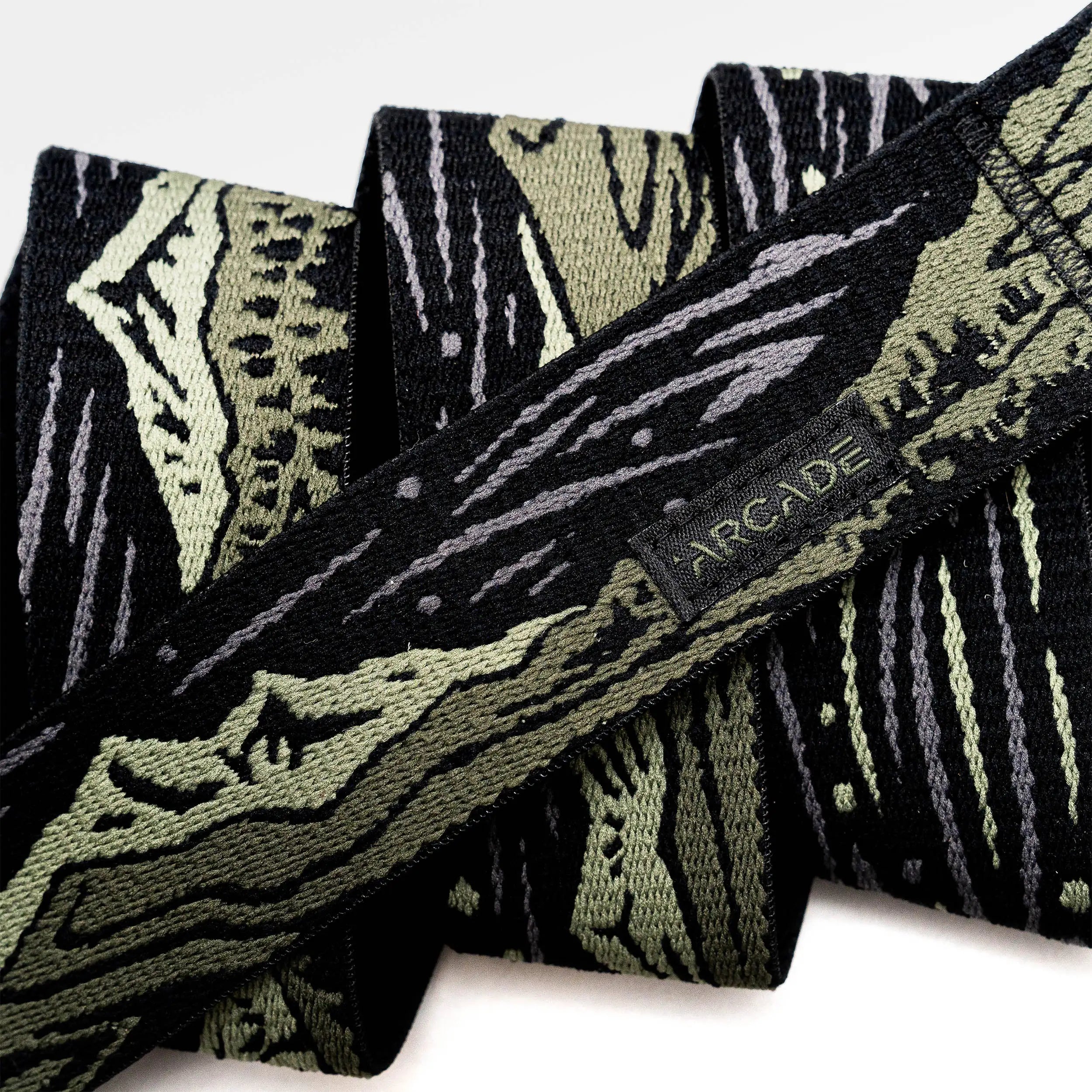 A close-up of the Arcade Belts Out of Range fabric belt, showcasing a black and olive green camo pattern, a small black tag with the brand name stitched on it, and a sturdy plastic buckle.