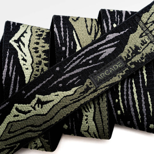 A close-up of the Arcade Belts Out of Range fabric belt, showcasing a black and olive green camo pattern, a small black tag with the brand name stitched on it, and a sturdy plastic buckle.
