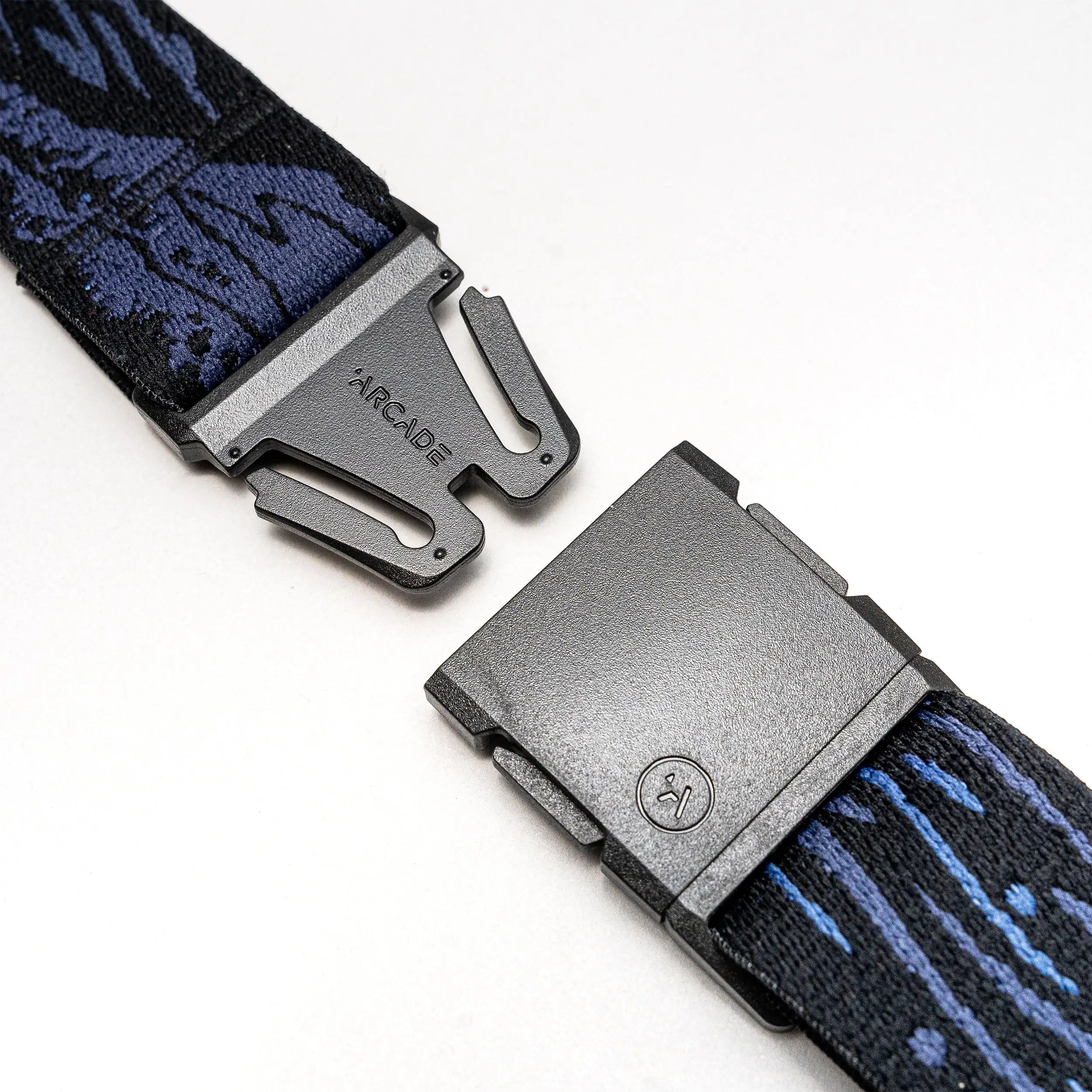 Close-up of the Arcade Belts Out of Range travel belt in black and blue pattern, featuring a metal quick-release buckle engraved with ARCADE and a small circular logo on one end. The belt is partially detached to highlight its low-profile design.