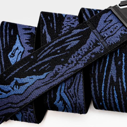 A close-up of the Arcade Belts Out of Range belt, showcasing its low profile design, black and blue abstract woodgrain pattern, and black buckle. The travel belt is neatly folded over itself multiple times.