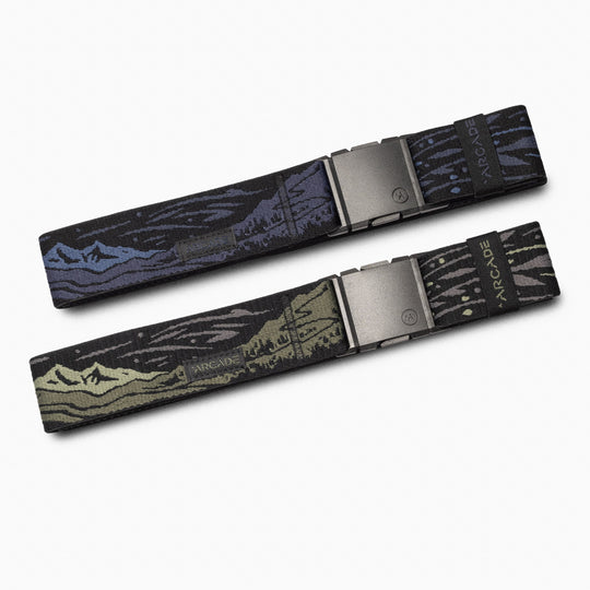 The Arcade Belts Out of Range Kit includes two elastic stretch travel belts with mountain and forest designs—one blue/purple, the other green/black—each featuring ARCADE branding on the metal buckle.