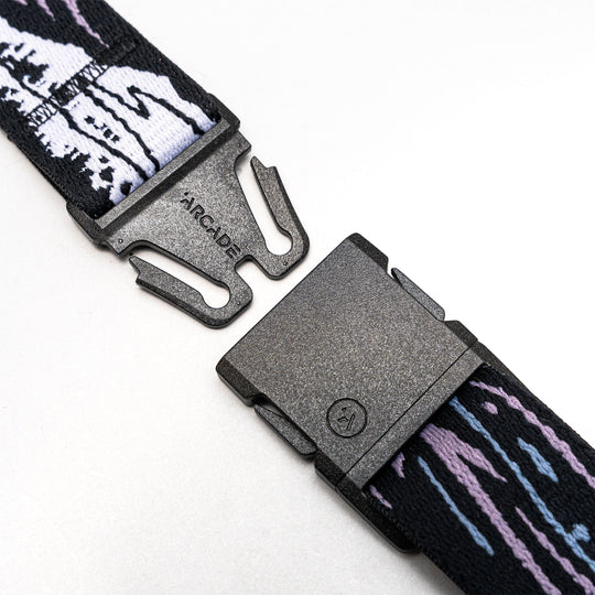 A close-up of the Arcade Belts Slim Kit shows its black plastic buckle on a stretch web strap with white, black, purple, and blue patterns, unclasped on a white background—perfect as a low-profile belt.