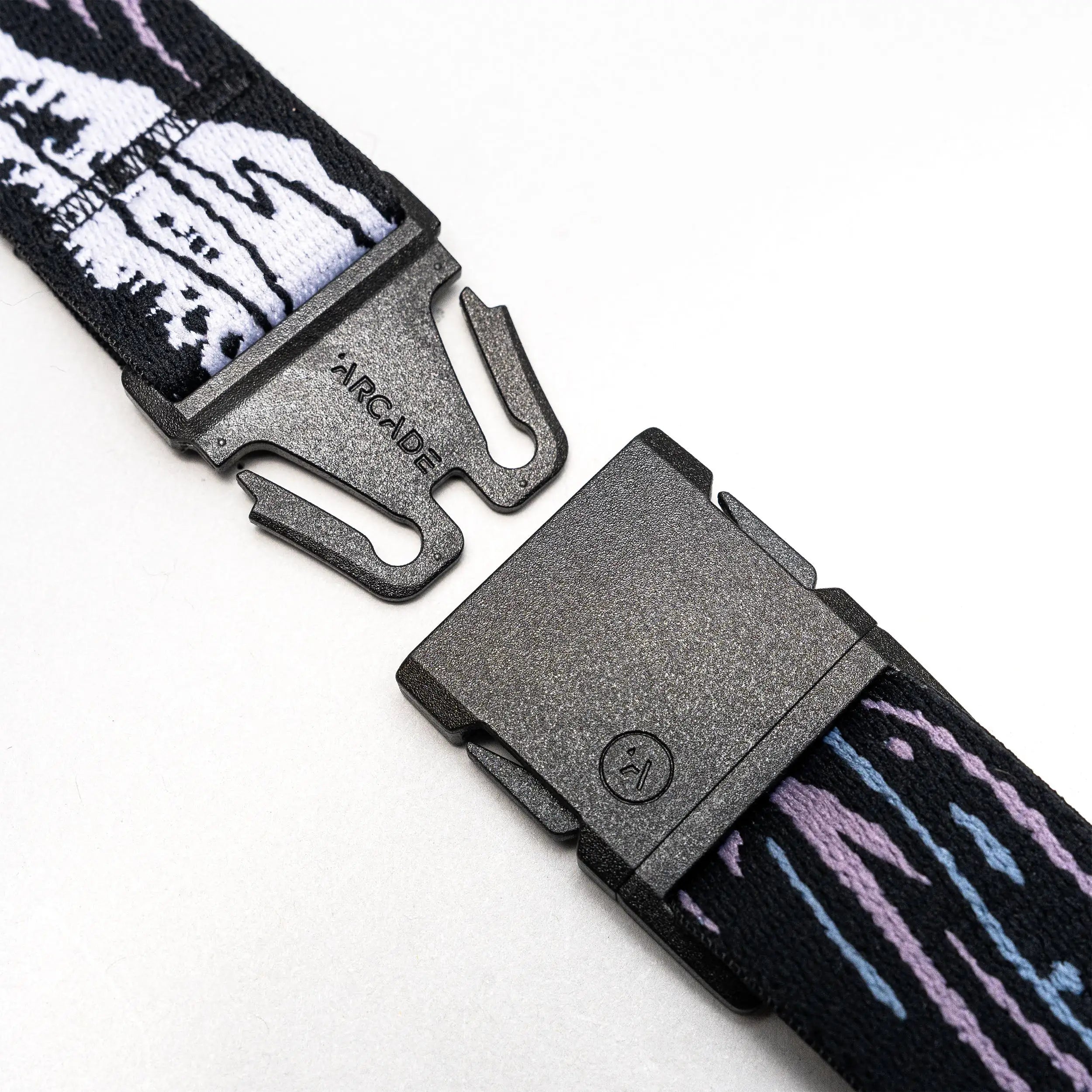 A close-up of the Arcade Belts Slim Kit shows its black plastic buckle on a stretch web strap with white, black, purple, and blue patterns, unclasped on a white background—perfect as a low-profile belt.