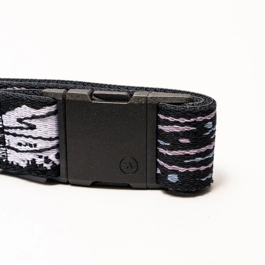 A close-up of the Arcade Belts Out of Range black elastic stretch belt with a black rectangular buckle and patterned fabric in black, white, and purple, coiled on a white background.