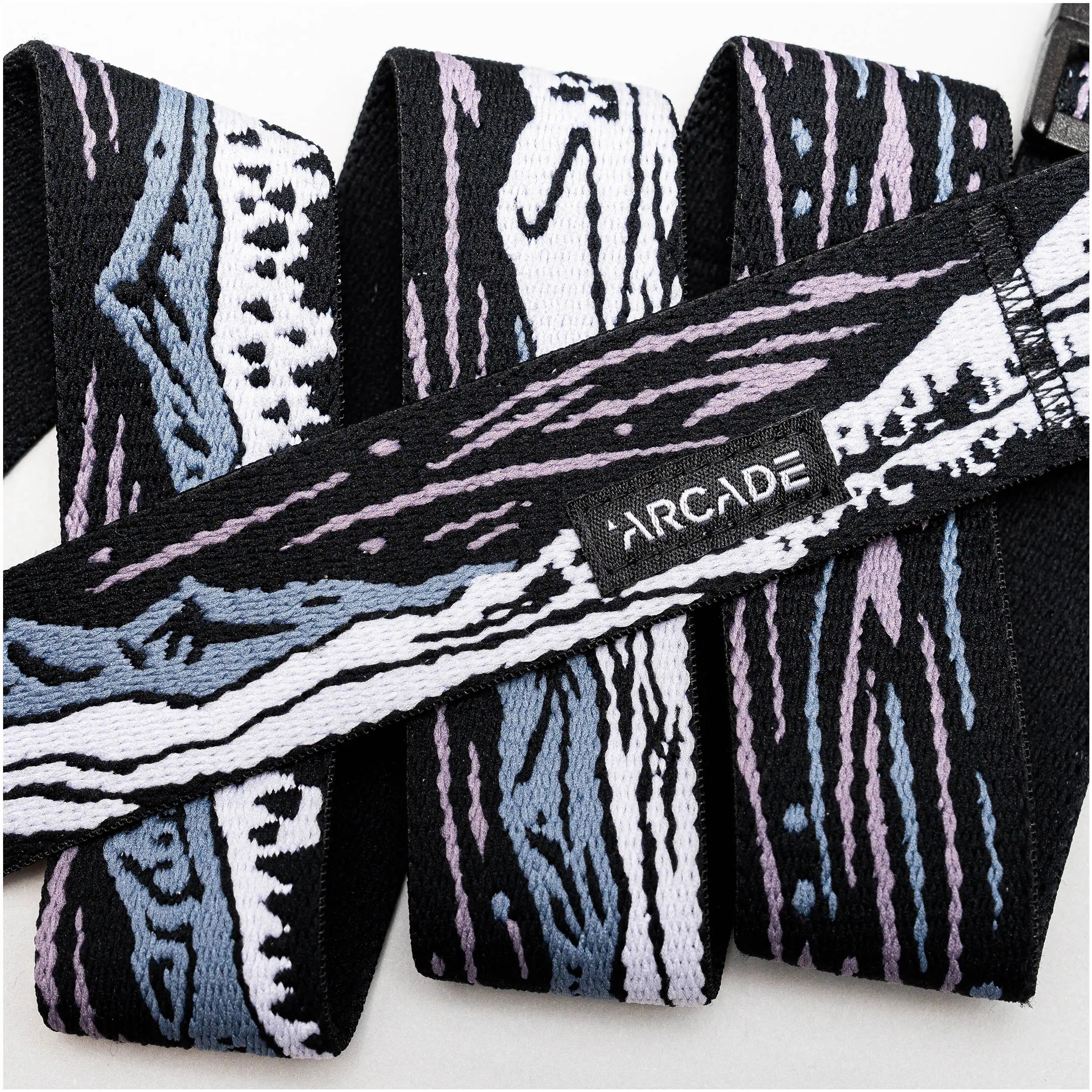 A close-up of the Arcade Belts Out of Range elastic stretch belt with an abstract black, white, blue, and lavender pattern, featuring a small black label stitched with white ARCADE text.