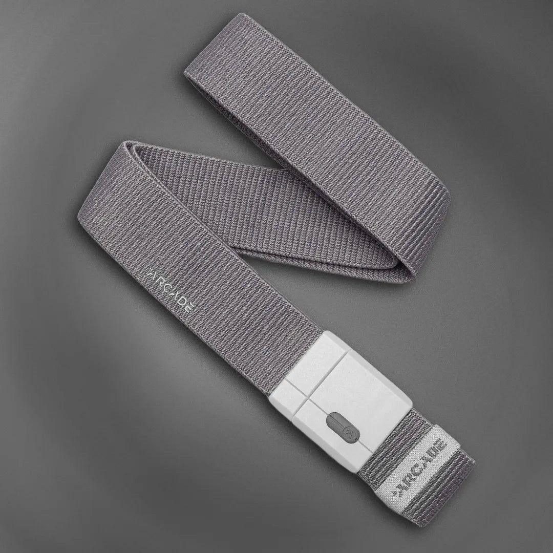 A gray, fabric belt with a white rectangular buckle lies coiled on a dark gray surface. The belt has the word ARCADE printed near the buckle.