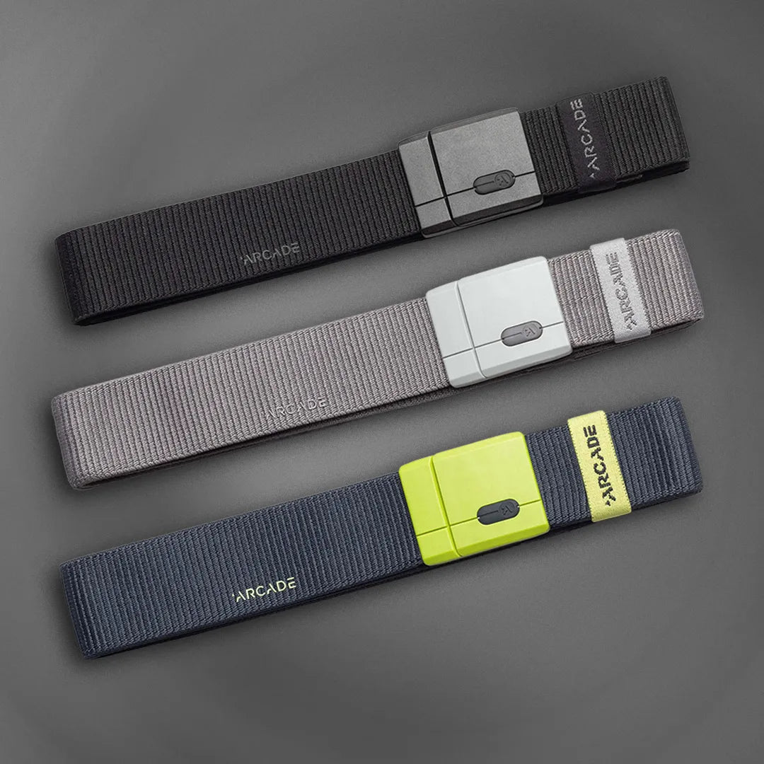 Three fabric belts laid parallel on a gray background: black with a dark gray buckle, gray with a light gray buckle, and navy blue with a neon yellow buckle, each labeled ARCADE near the end.