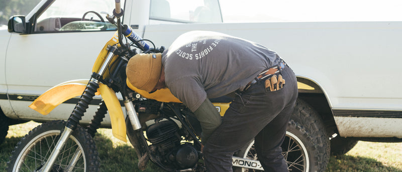 A person wearing a brown beanie and grey t-shirt, secured by a low profile belt, works on a yellow dirt bike parked next to a white pickup truck in an outdoor setting.