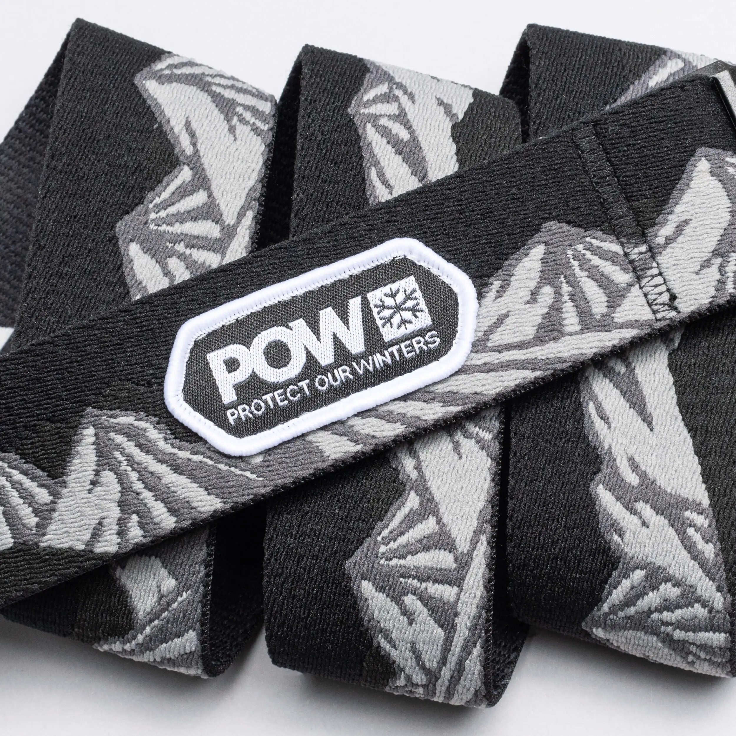 The Arcade Belts POW x Joseph Toney is a black and gray stretch webbing belt with mountain patterns, a low profile design, and a white POW PROTECT OUR WINTERS patch—showcasing classic Arcade Belts style.