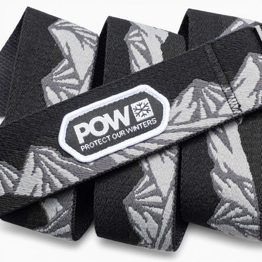 The Arcade Belts POW x Joseph Toney is a black and gray stretch webbing belt with mountain patterns, a low profile design, and a white POW PROTECT OUR WINTERS patch—showcasing classic Arcade Belts style.