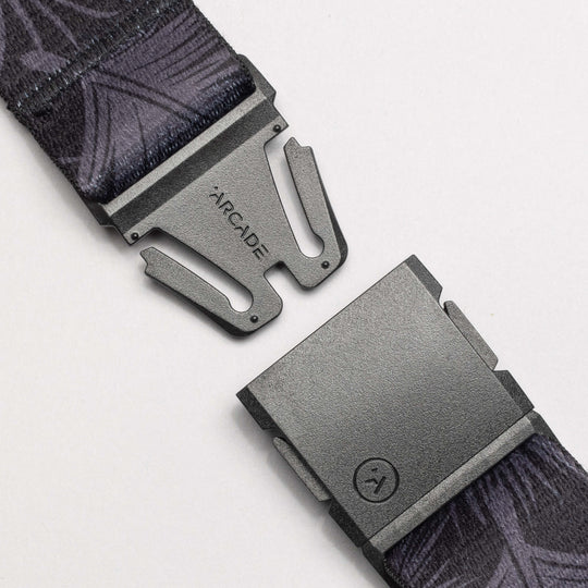 A close-up of a black geometric-patterned belt from Arcade Belts’ Island Collection, showing its gray metal buckle with ARCADE branding. The separated buckle ends rest on a light surface.