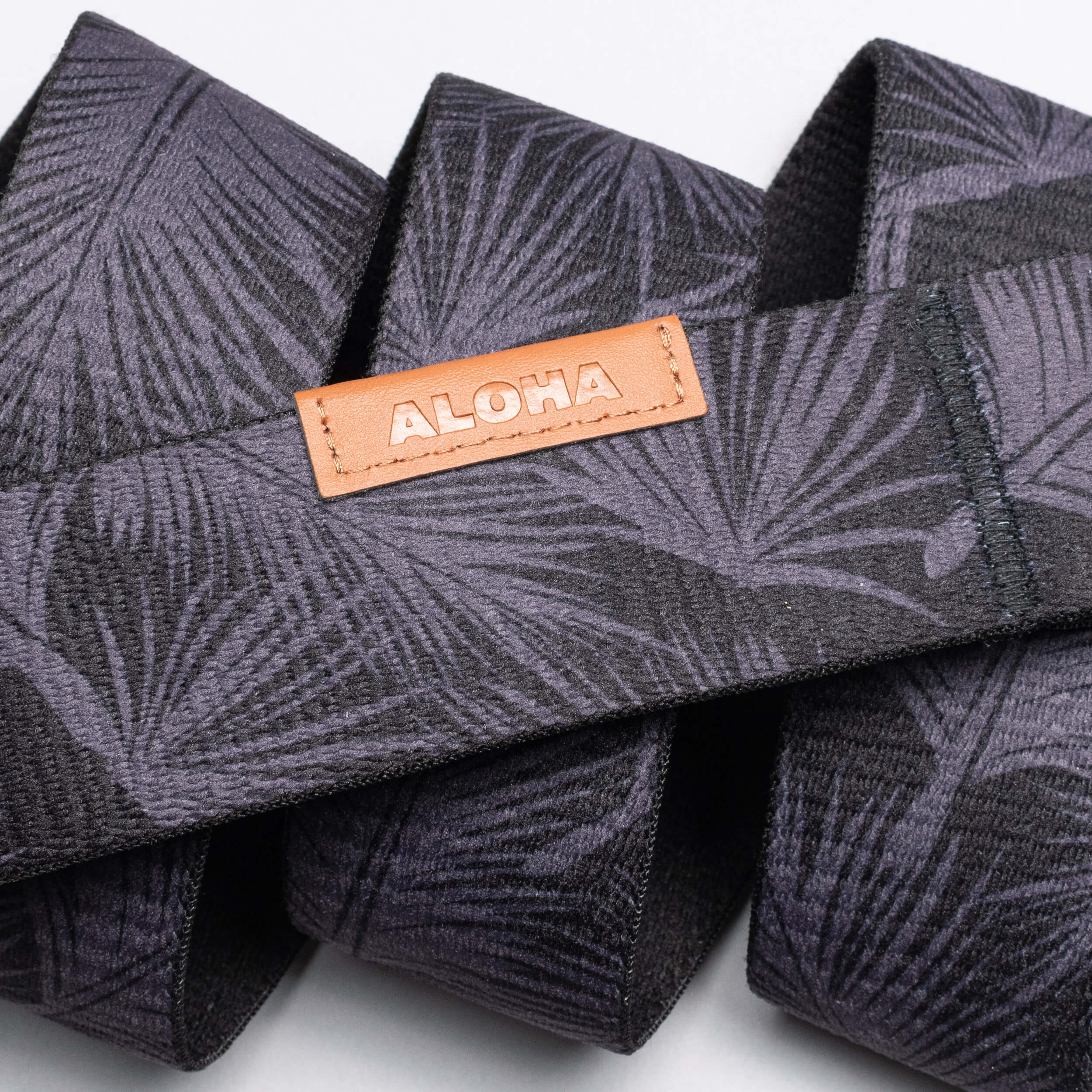 A close-up of a dark palm-leaf patterned Arcade Belts Island Collection band, featuring a tan leather patch embossed with ALOHA.