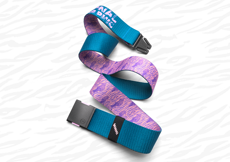 A fabric belt with a black buckle is shown on a white patterned surface. One side of the belt is teal blue, while the reverse side features a purple and pink abstract pattern. Text is partially visible near the buckle.