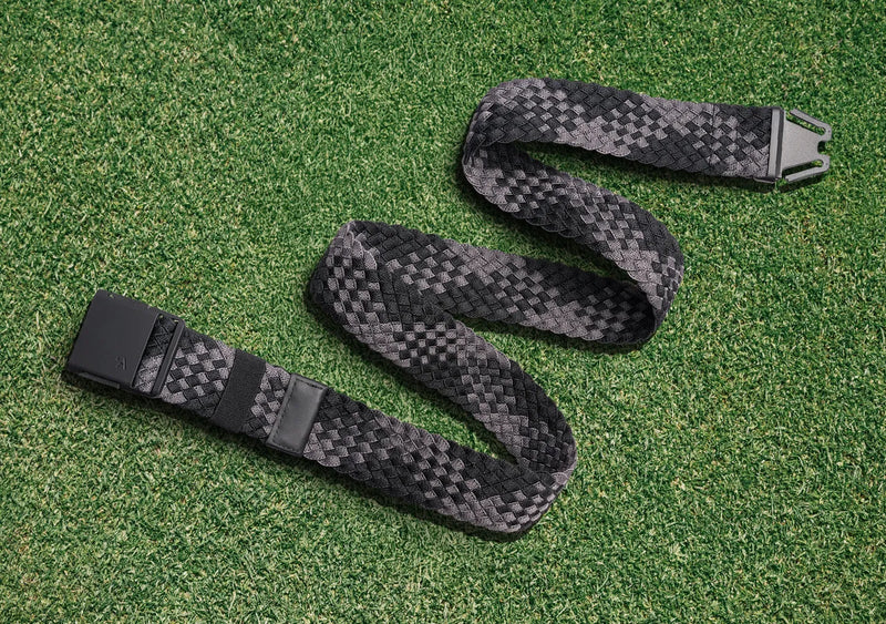 A woven black and gray belt with a black buckle is laid out in a zigzag shape on green grass.