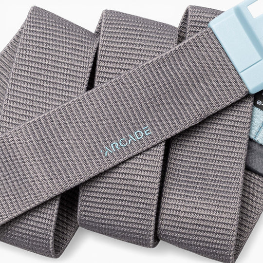 A gray ribbed fabric belt, the Polar Mag by Arcade Belts, is coiled against a white background. The word "ARCADE" is embroidered in light blue on its surface, and the durable light blue buckle features SwiftLock technology for easy, secure fastening.