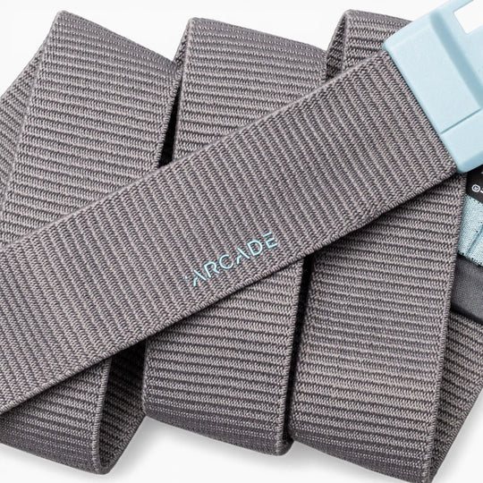 A gray ribbed fabric belt, the Polar Mag by Arcade Belts, is coiled against a white background. The word "ARCADE" is embroidered in light blue on its surface, and the durable light blue buckle features SwiftLock technology for easy, secure fastening.