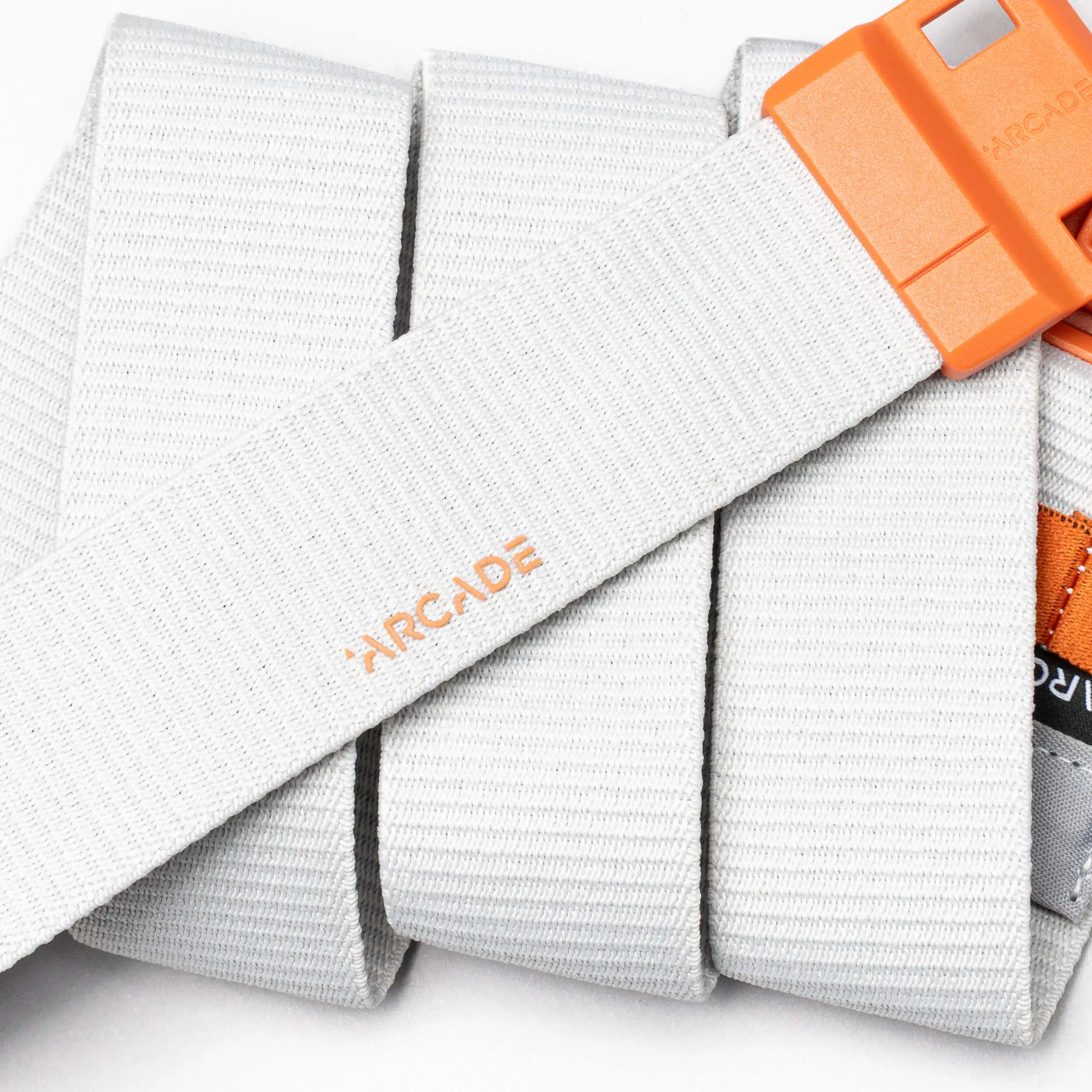 A close-up of the Arcade Belts Polar, a light gray belt with an orange SwiftLock buckle. The brand name "Arcade" appears in orange on its durable fabric, which is neatly folded in the background.