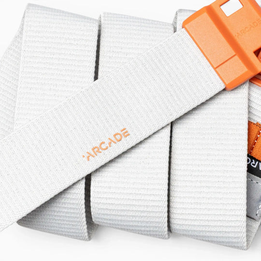 A close-up of a light gray Arcade Belts Polar Mag, a durable belt with an orange buckle, neatly rolled up. The Arcade logo is printed in orange on the belt, and it features SwiftLock technology for a secure fit.