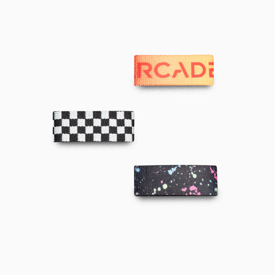 Three rectangular fabric patches—partial orange text on red, black and white checkerboard, and colorful paint splatter—Belt Loopkeepers by Arcade Belts in Pop color. Ideal accents or replacements for your favorite gear.