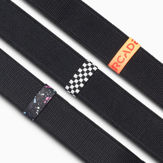 Three black Standard Width Arcade Belts are shown diagonally, each with a different Belt Loopkeeper: one with a space pattern, one checkerboard, and one featuring replacement elastic loops with an orange Arcade tag.