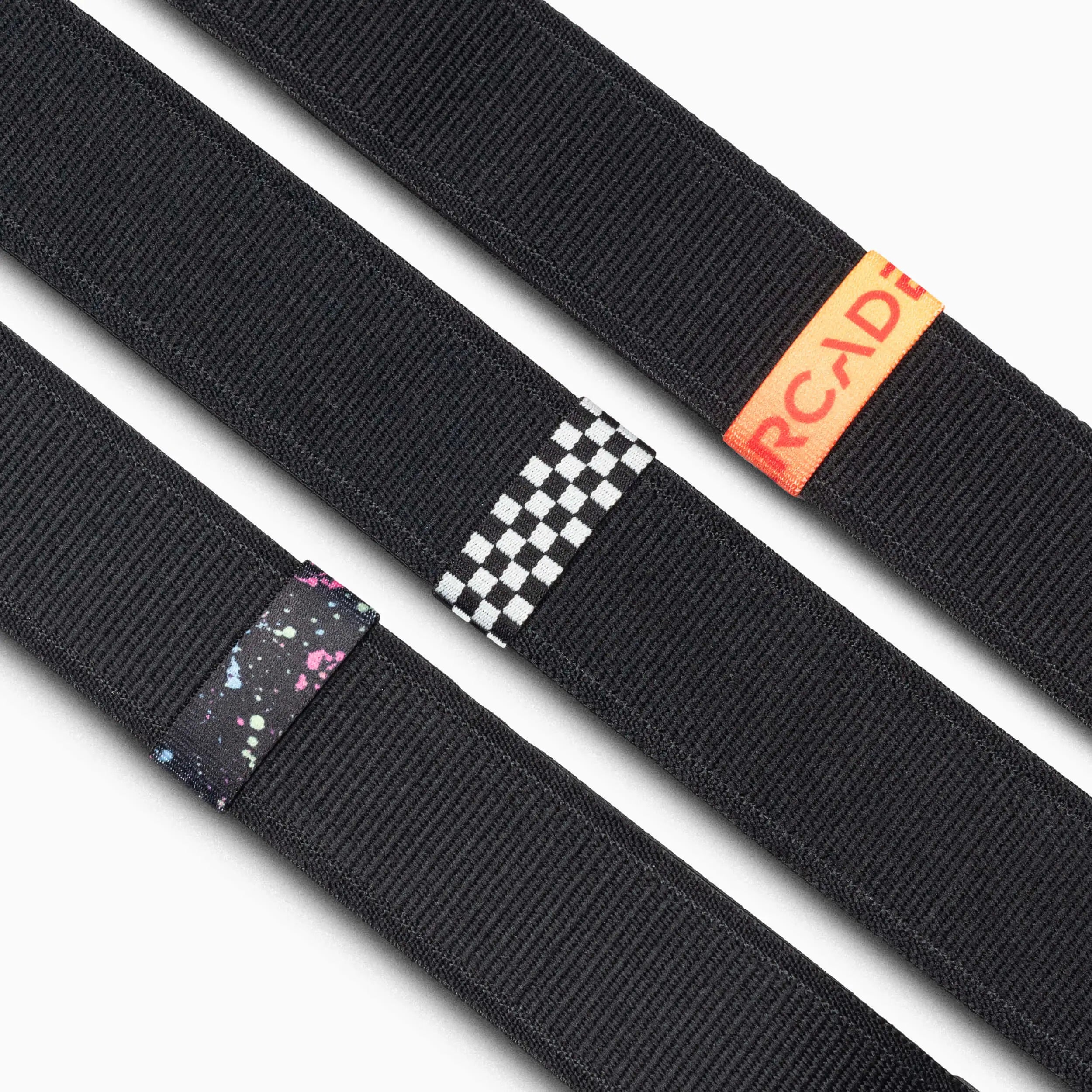 Three black Standard Width Arcade Belts are shown diagonally, each with a different Belt Loopkeeper: one with a space pattern, one checkerboard, and one featuring replacement elastic loops with an orange Arcade tag.