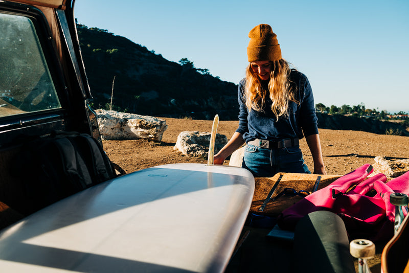 A woman wearing a brown beanie and blue sweater stands by an open car trunk, adjusting her Low Profile Belt with a Plastic Buckle as she looks down at a surfboard. The ocean and coastal cliffs shine in the sunlight behind her.