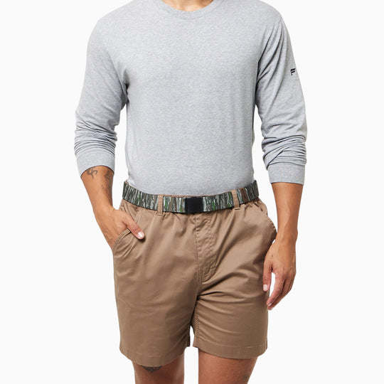A person stands with hands in pockets against a white background, wearing a light gray long-sleeve shirt, tan shorts, and the Arcade Belts Realtree Kit—a camouflage-patterned belt from Arcade Belts that’s an essential part of their camo gear.