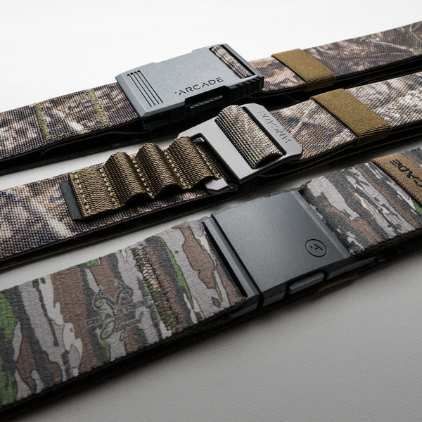 Three camouflage-patterned Arcade Belts with different black, metal, and plastic buckles are laid side by side on a white surface. The brand names Arcade and REALTREE EDGE are visible on the durable stretch webbing and buckles.