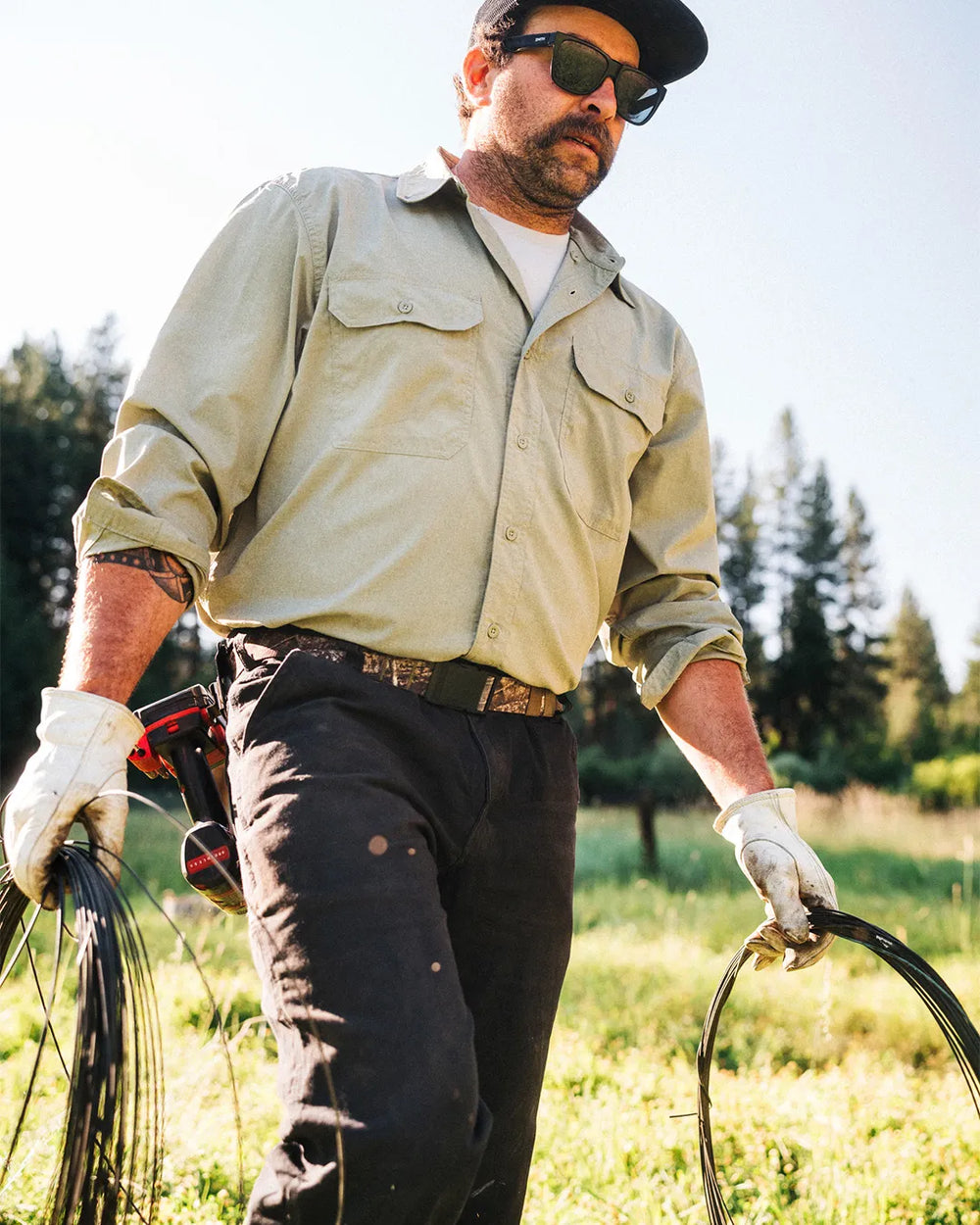 A man wearing sunglasses, a cap, gloves, and a light long-sleeve shirt stands outdoors in a grassy field holding coiled wire in both hands. Trees and sunlight appear in the background.