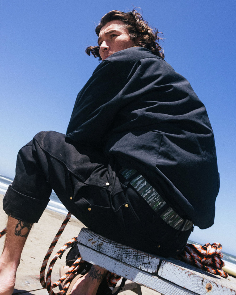 A person with long hair sits on a weathered wooden bench at the beach, dressed in dark clothing, looking over their shoulder. Coiled rope and a travel belt with a plastic buckle rest on the bench beneath the bright, clear sky.