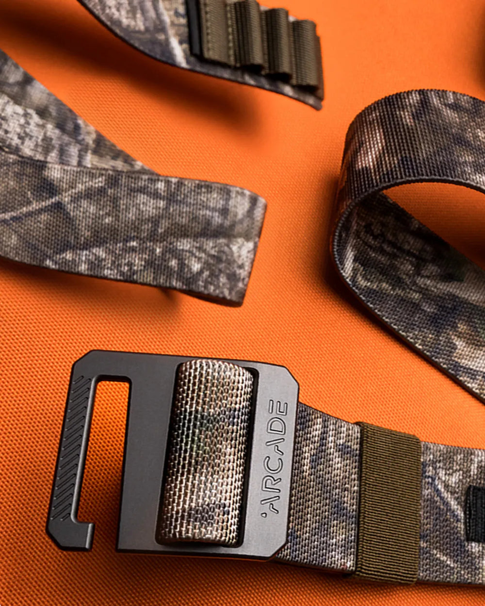 A camouflage-patterned belt with a metal buckle labeled ARCADE is displayed on an orange textured surface. The belt is partially coiled, showing both the buckle and the webbing.
