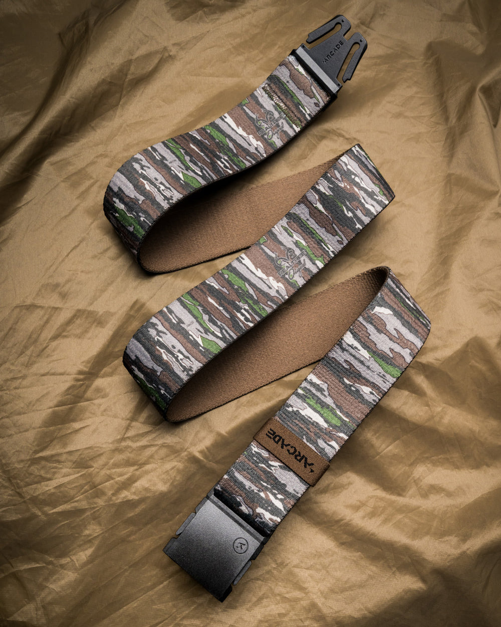 A camo-patterned Low Profile Belt with a black buckle lies on a crinkled tan surface. The brand Arcade is visible on a brown tag and buckle, showcasing the signature style of Arcade Belts.